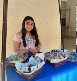 jovem rapariga a sorrir, em escola, numa banquinha com toalha azul e vários cestos com artigos solidários Make-A-Wish